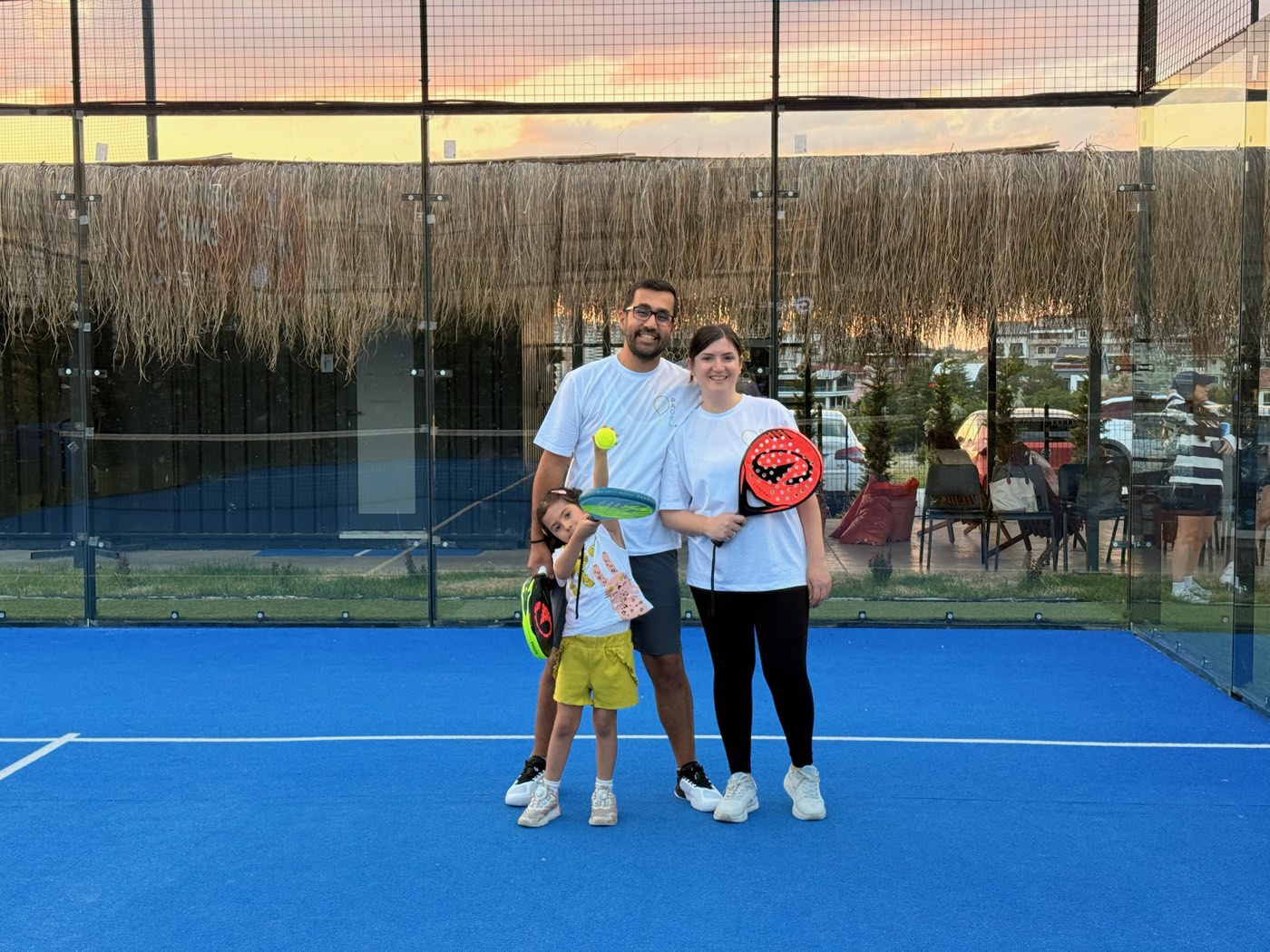Padel kortunda raketleriyle poz veren anne, baba ve kızları — gün batımında, mavi zeminli kortta